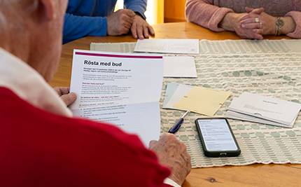 En man håller ett brev med information om budröstning, mobiltelefon och valsedlar bredvid. Två personer till vid bordet.