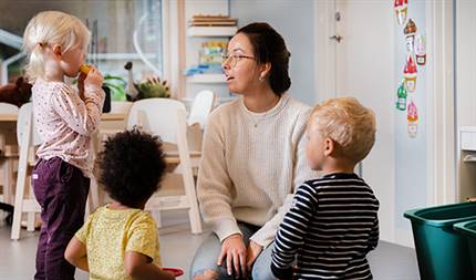 Ung kvinna sitter på golvet och pratar med tre förskolebarn